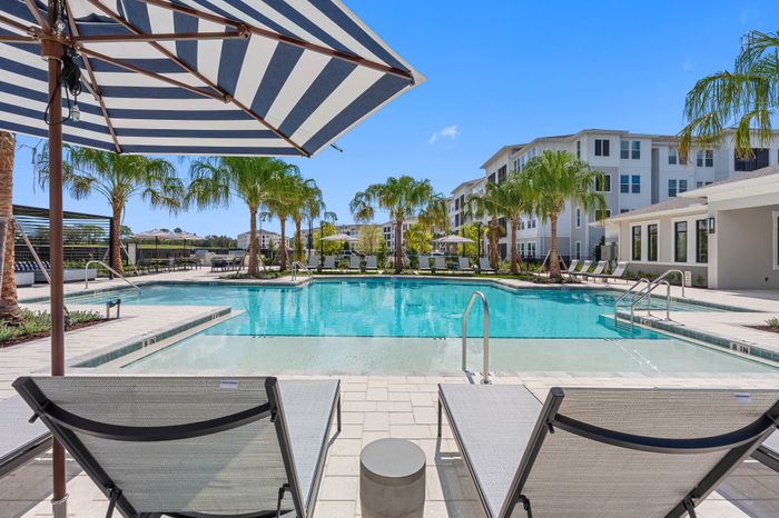 Luxurious apartment complex with a pristine swimming pool bordered by palm trees and modern sun loungers, under a clear blue sky.