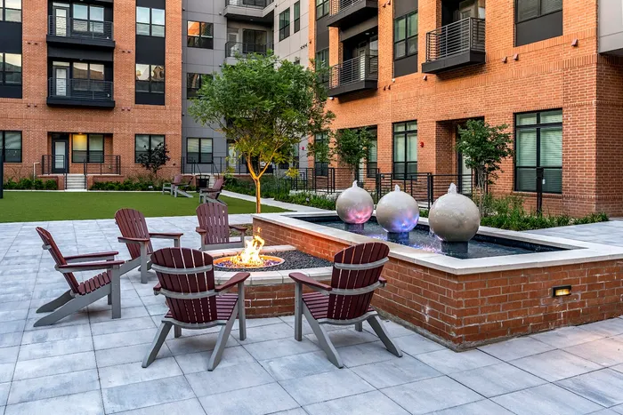 Charming apartment courtyard featuring cozy Adirondack chairs around a glowing fire pit, elegant water features, and lush greenery.