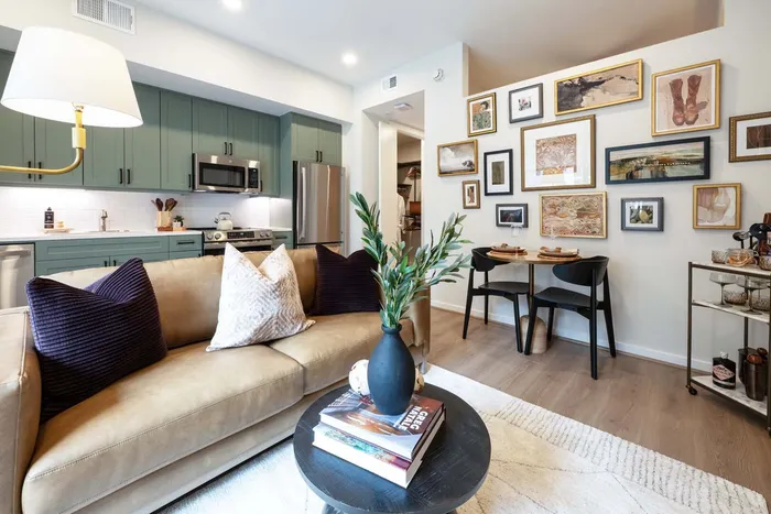 Charming open-plan living space featuring a cozy sofa with plush cushions, stylish coffee table with books, and modern kitchen with sage green cabinets. Beautifully decorated dining area with art-adorned wall and elegant dining set.