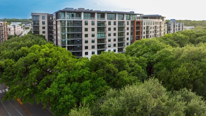 Modern apartment building surrounded by lush green trees, featuring expansive glass windows and a sleek, contemporary design.