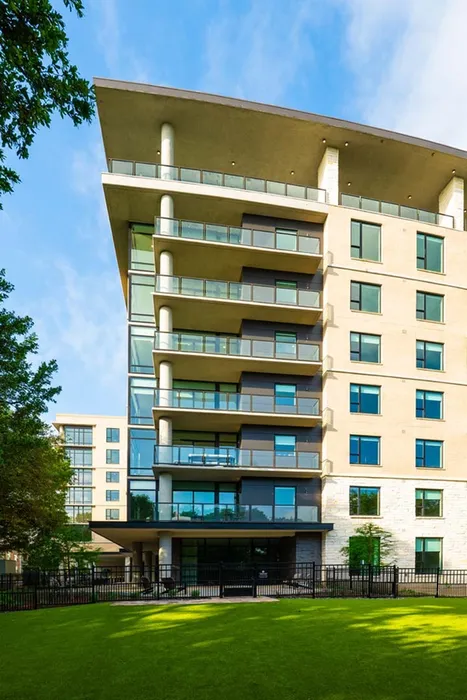 Modern apartment building with expansive balconies and sleek glass railings, surrounded by lush greenery and clear blue skies.