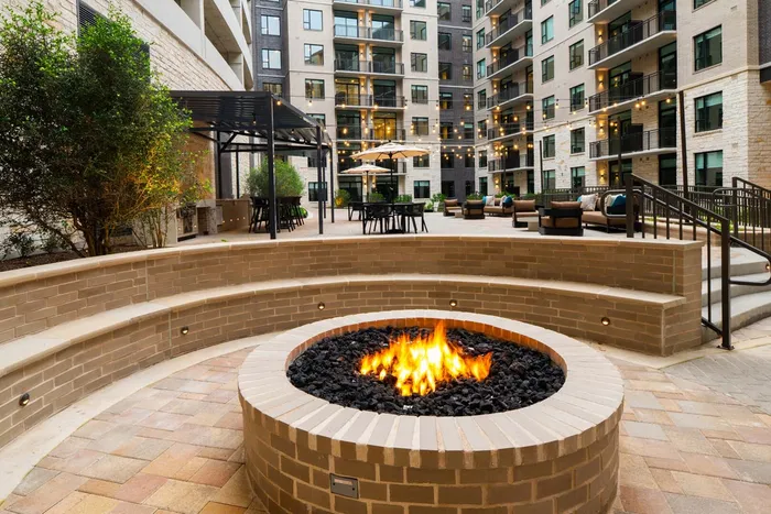 Modern apartment courtyard featuring a cozy fire pit, elegant seating areas, string lights, and lush greenery for urban outdoor relaxation.