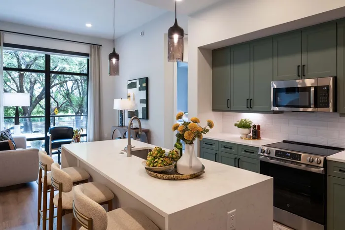 Bright modern kitchen with island seating, stylish green cabinets, and a spacious open-concept living area with large windows and natural light.