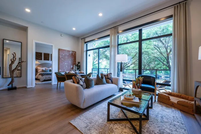 Bright living room with elegant decor, featuring a cozy sofa, large windows overlooking greenery, and access to a beautifully furnished bedroom.