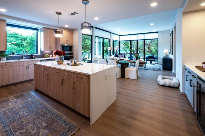Luxurious open-plan kitchen and living area with modern wooden cabinetry, elegant island, and floor-to-ceiling windows offering lush greenery views.