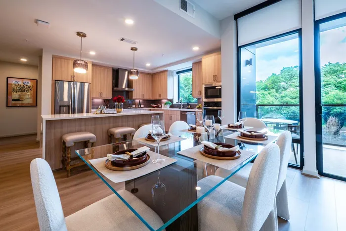 Elegant contemporary dining area with glass table and plush chairs, adjacent to a modern kitchen boasting sleek cabinetry and stainless steel appliances, illuminated by natural light through large windows.