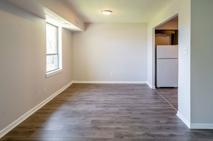 Empty living room with gray wood-look flooring, window and kitchen entry at Woodcrest Village Apartments in Fairless Hills.