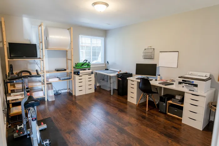 Multi-use workspace with desk, printer, shelving, and exercise bike, a window, and hardwood-style flooring.