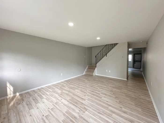 Woodland Manor living room: light wood floors, gray walls, ceiling lights, stair with metal railing and hallway to kitchen.