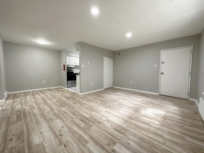 Spacious living area with wood-look flooring, gray walls, kitchen opening, and entry door at Woodland Manor.