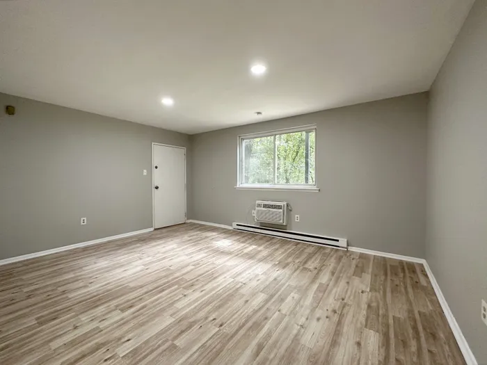Spacious empty living room with wood-look floors, gray walls, wall air conditioning unit and entry door at Woodland Manor.