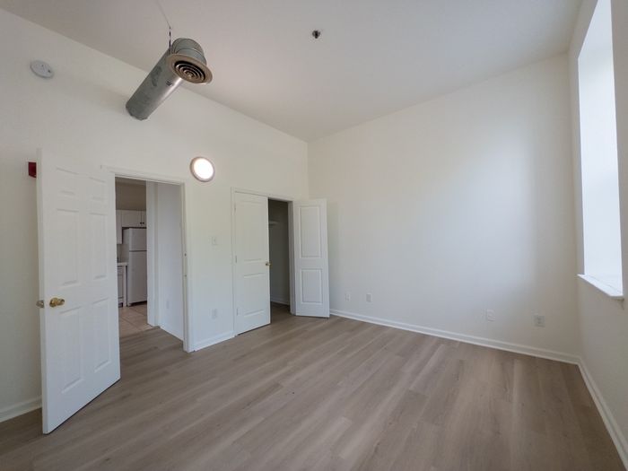 spacious bedroom with exposed air vent and vinyl plank flooring, tall window, loft style apartment, deep closet and door leading towards kitchen
