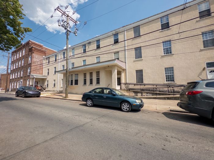 long concrete building with lots of windows attached to tall brick building, sidewalk linking streets, cars parked along street