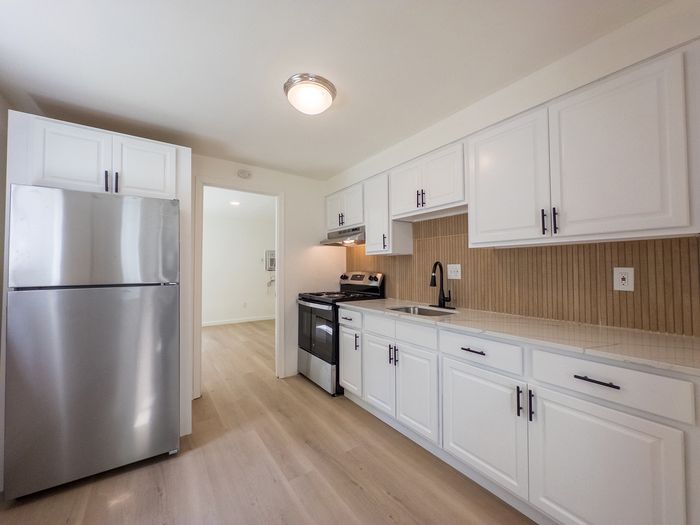 kitchen with white cabinets, stainless steel fridge and oven, slatted tile backsplash and overhead light, door leading to bedroom