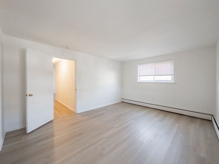 apartment bedroom with vinyl plank flooring, bright window with blinds, door leading to well-lit hallway