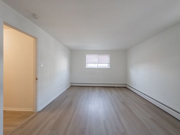 apartment bedroom with vinyl wood floor, bright window, door leading to hallway, baseboard heating