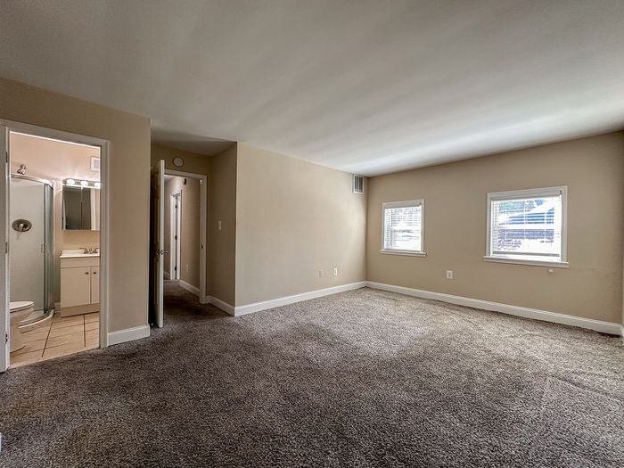 spacious carpeted bedroom with attached full bathroom, bright windows