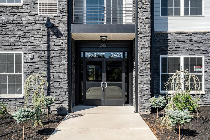 Chic apartment entrance with modern stone facade and lush landscaping at The Warrington Place, showcasing stylish urban living.