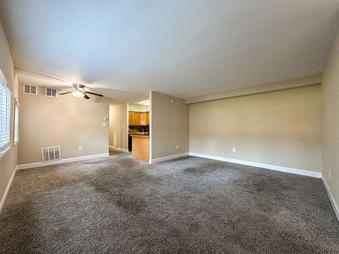 Spacious living area with plush carpeting, ceiling fan, and open view to the modern kitchen with wooden cabinets, featuring ample natural light.