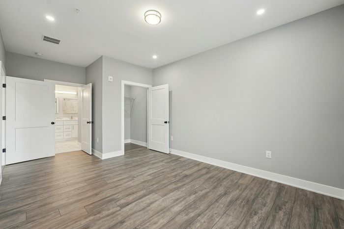 Modern empty room with elegant wood flooring, fresh gray walls, and white doors leading to a stylish bathroom and closet. Abundant natural light creates an inviting atmosphere.