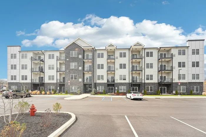 Modern four-story apartment building with balconies, set against a bright blue sky with scattered clouds and featuring ample parking space.