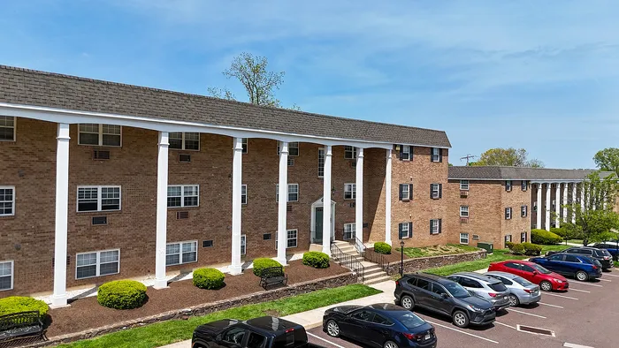 Charming brick apartment building with elegant white columns and well-maintained landscaping, showcasing ample parking spaces under a clear blue sky.