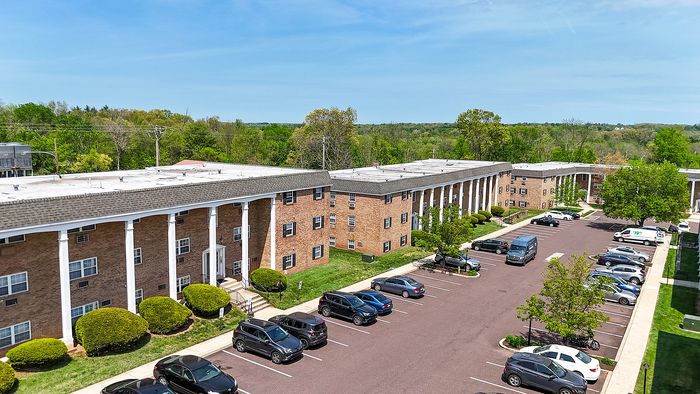 Charming apartment complex with classic brick facade, expansive parking space, and lush green surroundings, offering a peaceful residential environment.