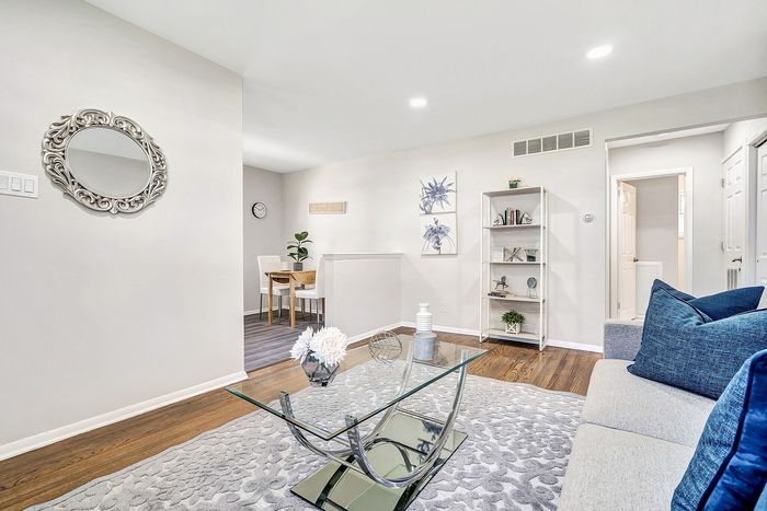Modern living area with a chic glass coffee table, plush gray sofa, elegant wall mirror, and sleek decor, leading to a cozy dining nook with stylish furniture.