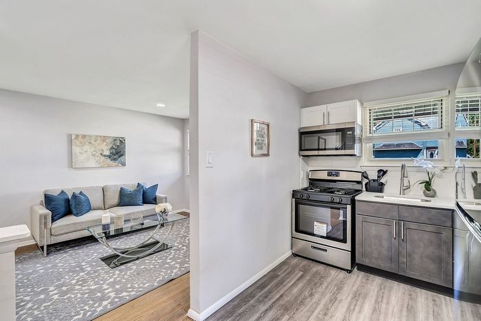 Modern open-concept living and kitchen area featuring a cozy sofa with blue accent pillows and a sleek glass coffee table, adjacent to a stylish kitchen with stainless steel appliances and abundant natural light.
