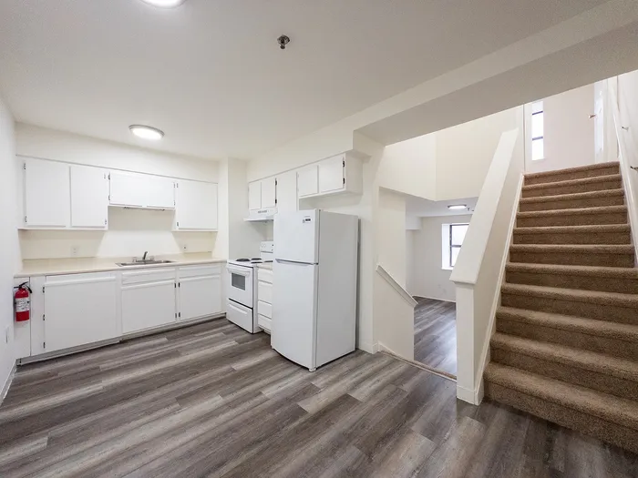 Modern open-plan kitchen with white cabinetry, sleek appliances, and elegant wood flooring adjacent to a carpeted staircase leading to a bright living area.