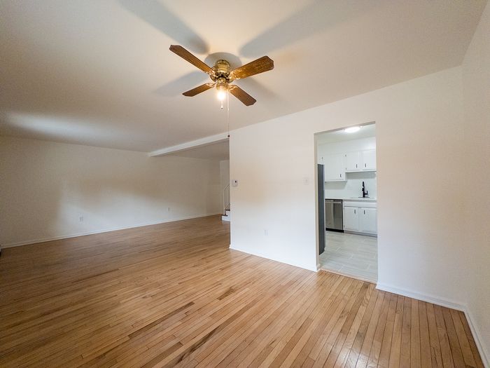 Spacious open living room with gleaming wooden floors, ceiling fan, and a view into a modern, well-lit kitchen.