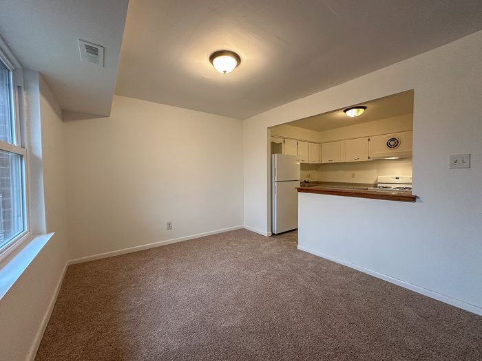 Carpeted living room with window and half-wall opening to a kitchen with white appliances at Woodcrest Village.
