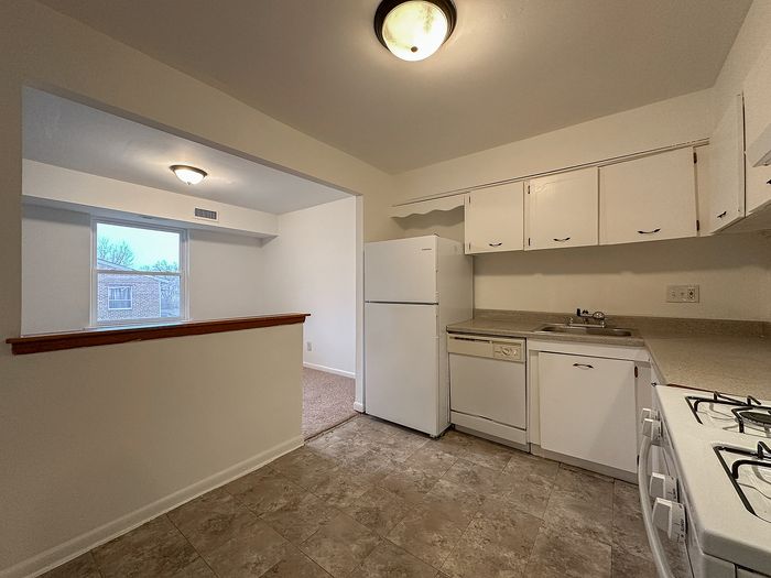All-white corner kitchen with refrigerator, dishwasher, gas range, overhead cabinets, and tile floor at Woodcrest Village.
