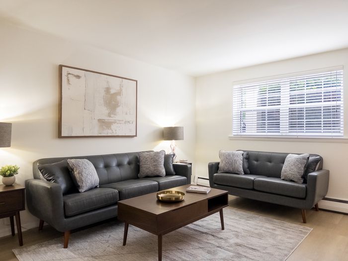 Furnished living room at College Arms Apartments with two gray sofas, coffee table, wall art, and window blinds.