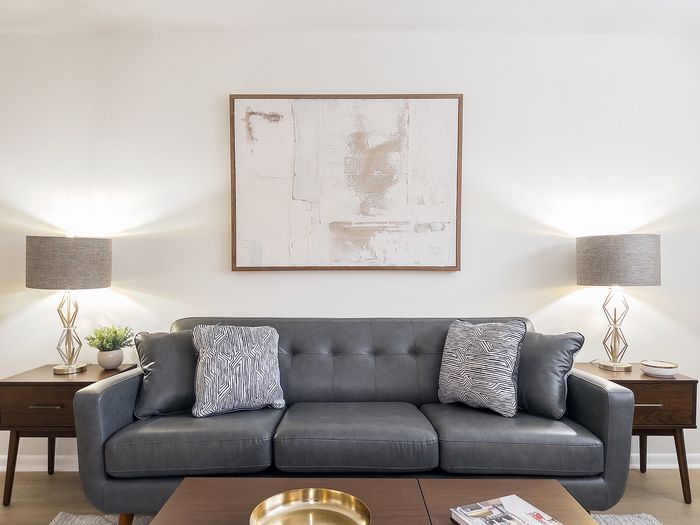Living room with gray leather sofa, abstract wall art, and lamps at College Arms Apartments.