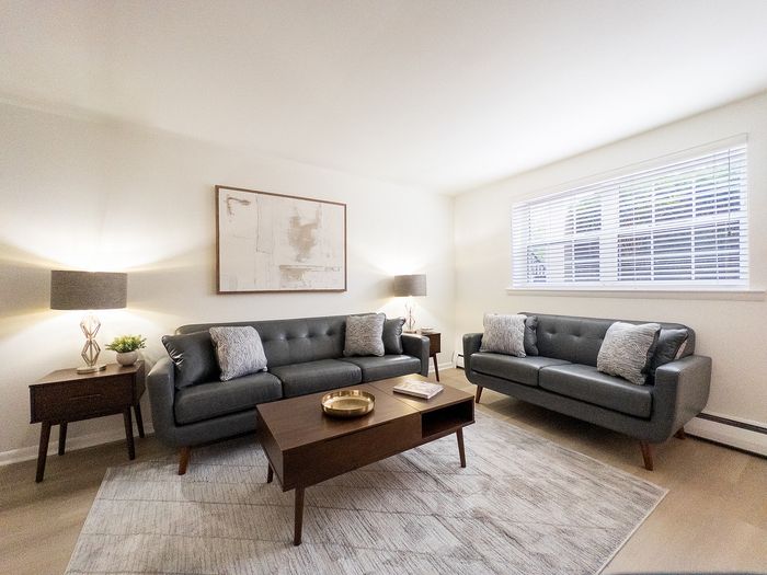 Living room at College Arms Apartments with two gray sofas, wooden coffee table, lamps, rug, and blinds.