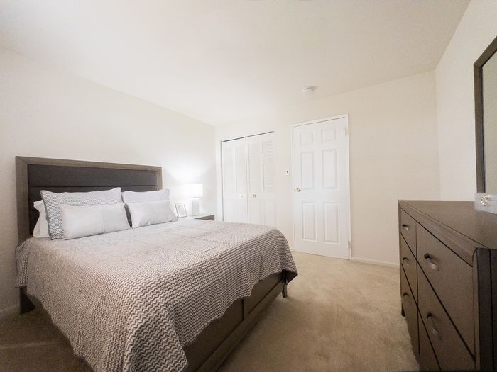 A bedroom with queen bed, nightstand, dresser, and carpeted floors at College Arms Apartments.