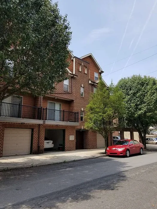 Charming brick townhouse with lush green trees, featuring a two-car garage and a cozy balcony.