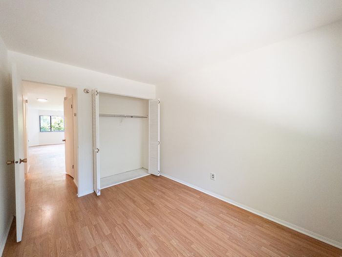 Bright, spacious bedroom with wood flooring and open closet, leading to a sunlit living area with garden view.