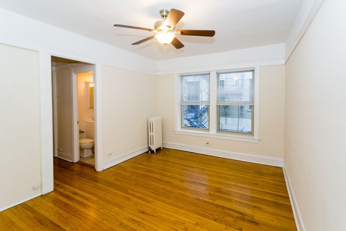 A living room with hardwood floors, two sunny windows, a ceiling fan, and off white walls.
