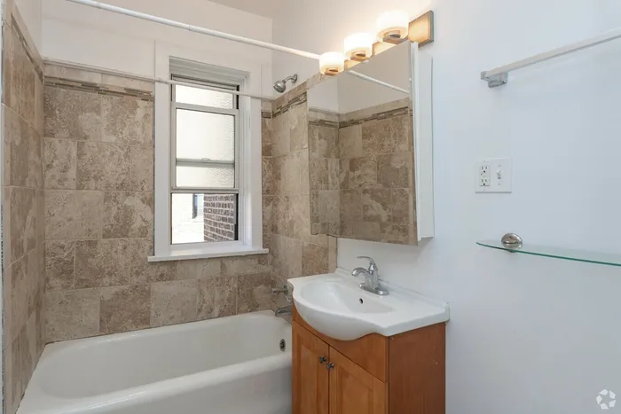 A bathroom with a medicine cabinet, wooden vanity, and stone tile tub surround.