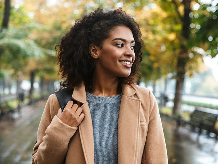 A smiling prospective female resident with dark curly hair and a camel hair coat walking in Hyde Park.
