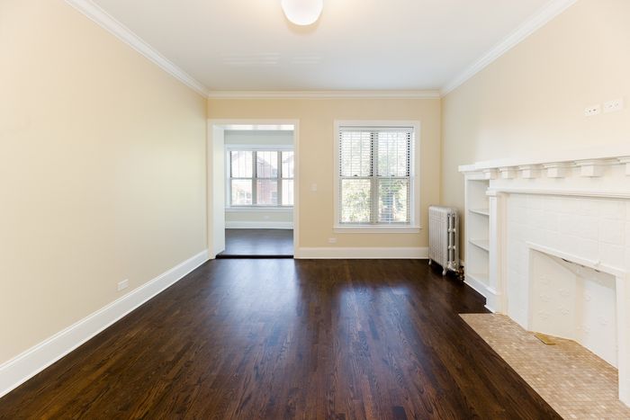 A living room with dark hardwood floors, sunny windows, a decorative fireplace, and off white walls with white trim.