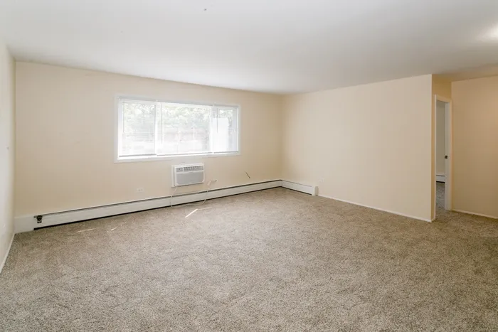 Bright and sunny living room with wall to all beige carpeting and a sleeve air conditioner.