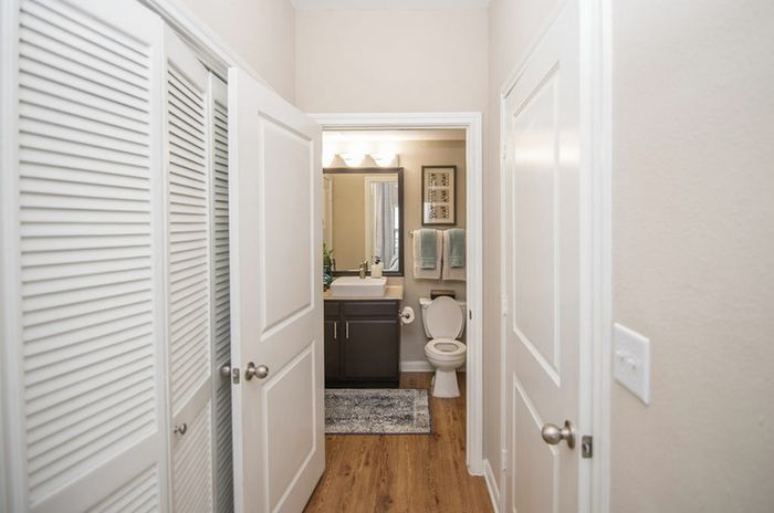 a hallways with white walls and a hardwood floor, leading to a modern bathroom with a large mirror, dark cabinets, and a white sink