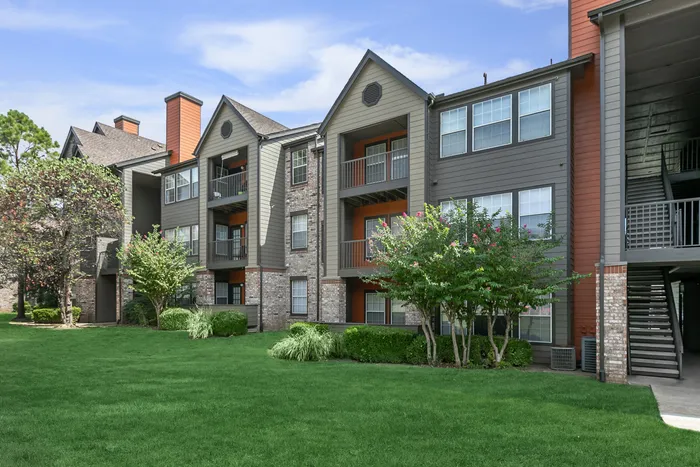 the outside view of a three-floor apartment building surrounded by a vibrant lawn of grass, bushes, and trees