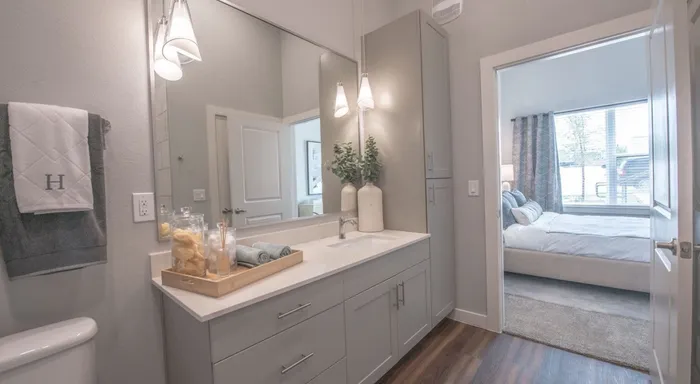 modern bathroom with light walls, a white countertop and sink, a vase with a plant, a tray with towels and sponges, two towels, hardwood flooring, and a large mirror
