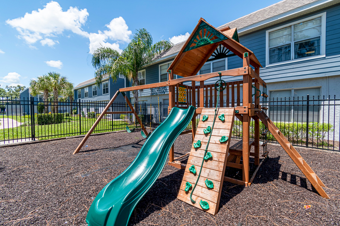 Playground with slide, swings, and climbing wall, surrounded by a fence.