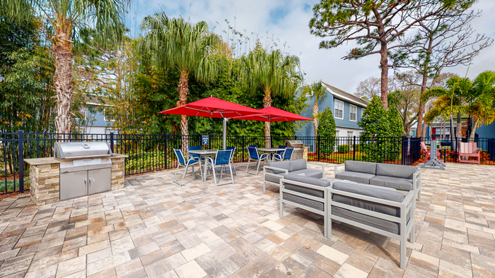 Pool grill area with cushioned gray patio furniture, gray table and blue chairs, red umbrellas, trees, blue apartments