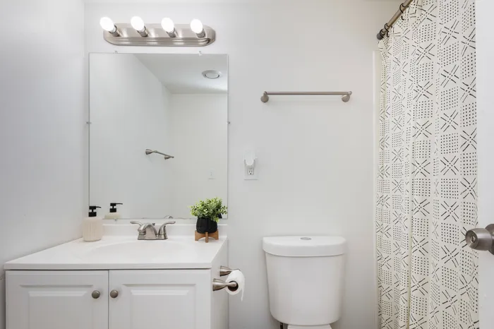 Spacious bathroom with white cabinetry, contemporary lighting above vanity, decorated with plants and patterned shower curtain